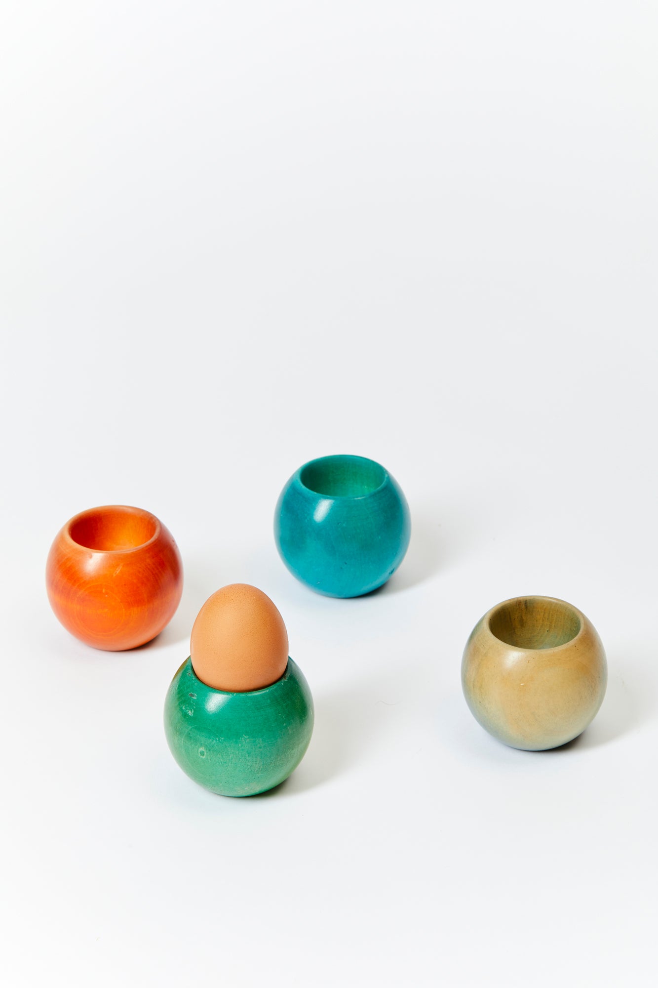 Four colored wooden egg cups arranged on a white background, one holding a brown egg. Each cup is a different color: orange, green, blue, and natural. 