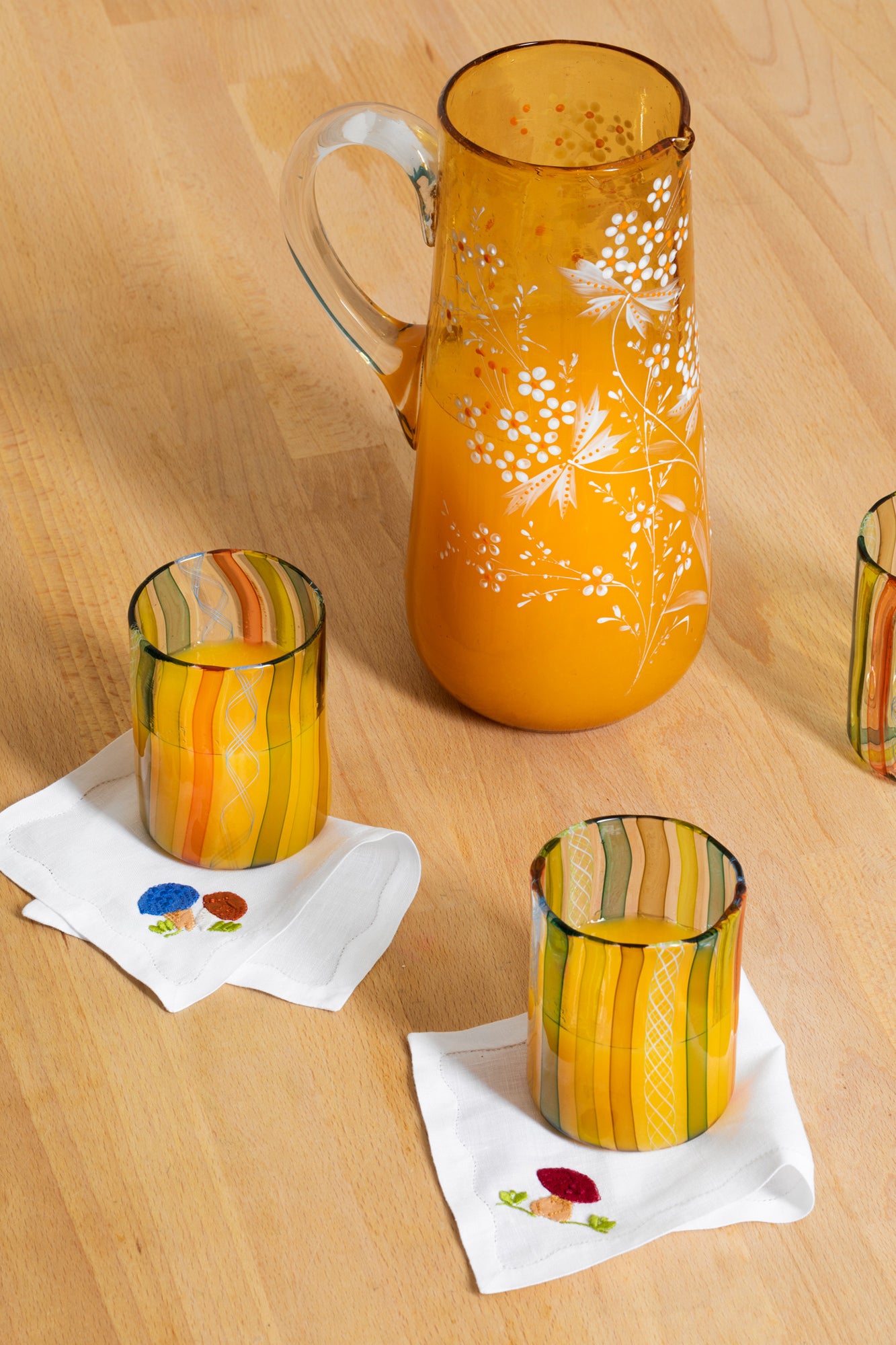Photograph of table setting featuring hand blown glass tumblers atop French stitch appliqué cocktail napkins, next to a vintage glass pitcher. 