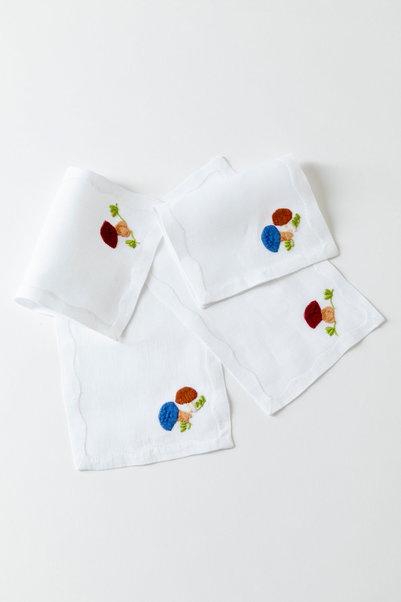 Cocktail napkins with French stitch appliqué made in Madeira, Portugal. 4 napkins against a white background with 2 different designs. Both napkins are white with an embroidery in the corner. One is of a mushroom with a maroon cap and some green grass sprigs. The other design has two mushrooms, one with a blue cap and one with a red cap, and some green grass sprigs.