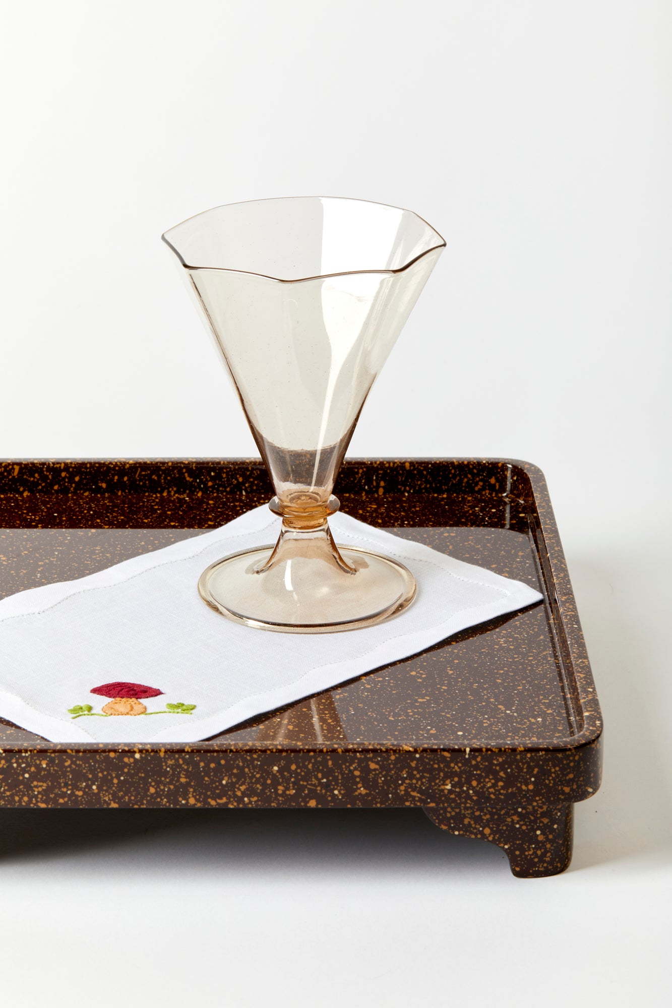 Photograph of a hand blown glass goblet resting on an embroidered cocktail napkin on top of a serving tray. 
