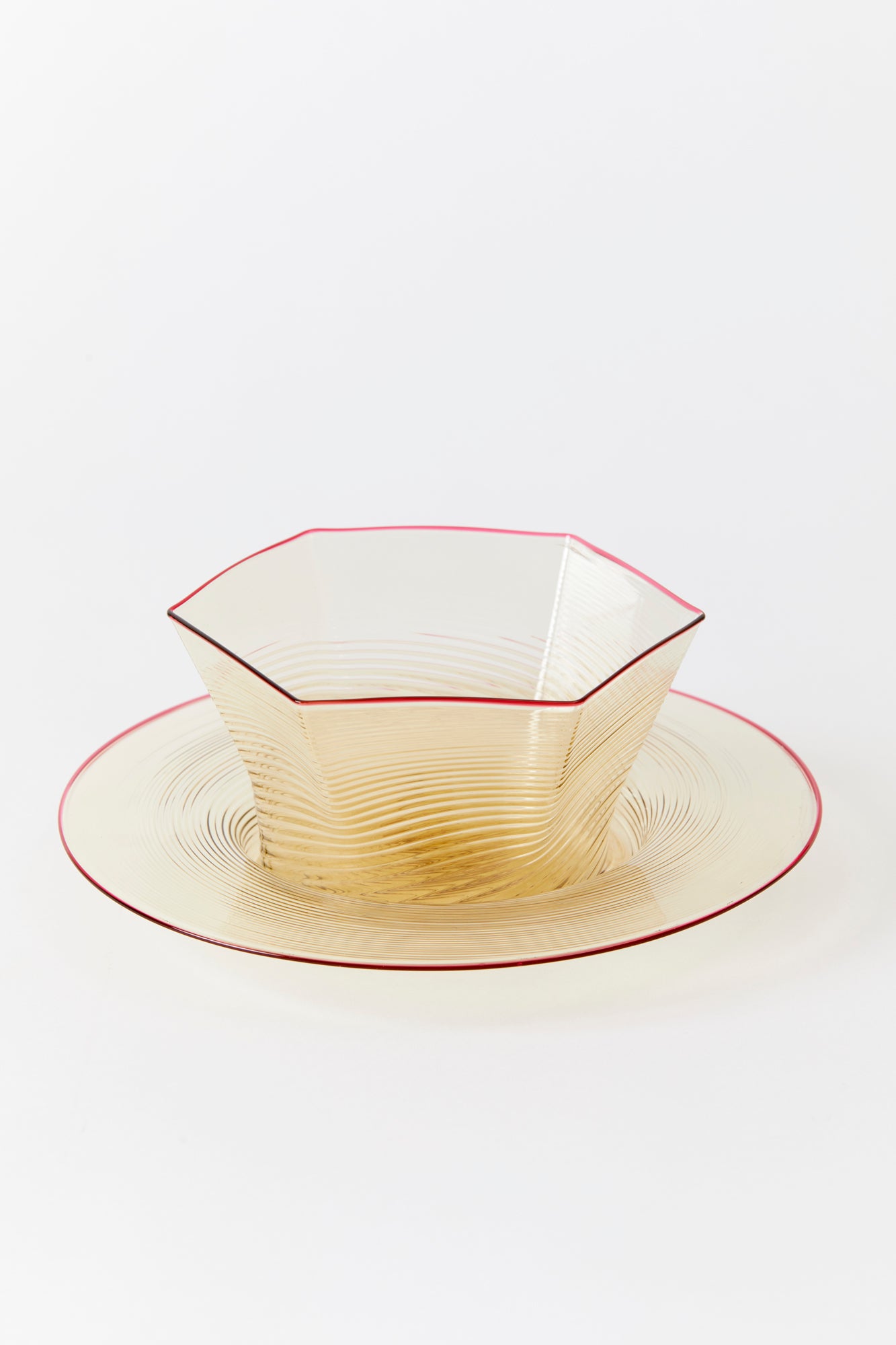 Glass blown and saucer on white background. Both pieces are hand blown in a warm smoky colored glass with a red rim and swirling pattern. The bowl has a hexagonal shape and sits inside the wide saucer. 