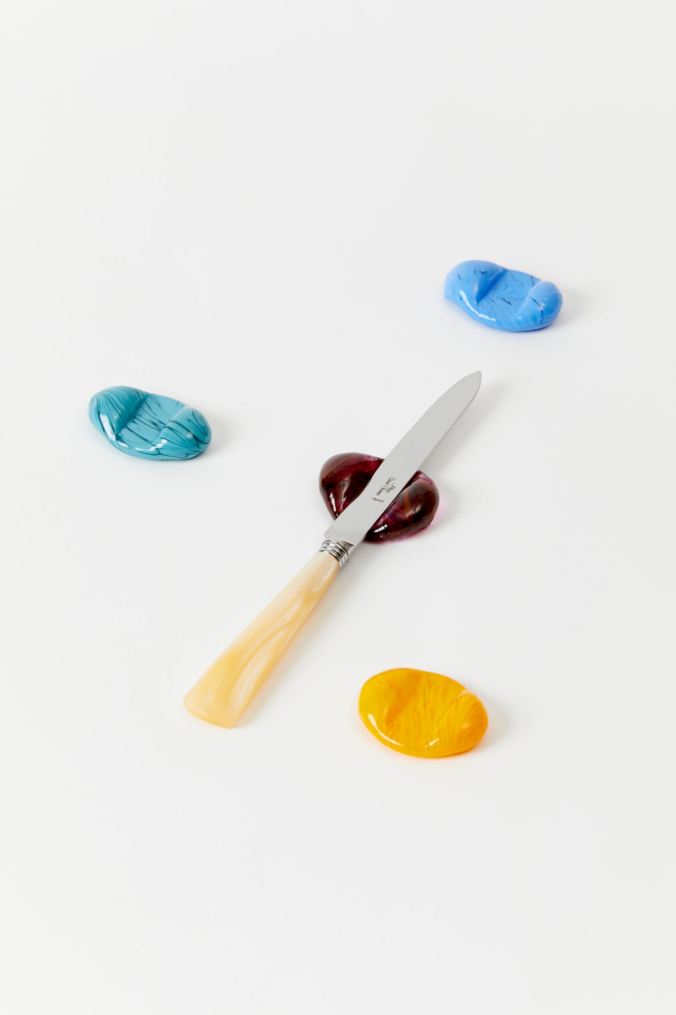 4 hand blown glass knife rests on a white background, 1 with a mother of pearl knife atop. Knife rests are all different colors: teal, blue, maroon, and yellow. 