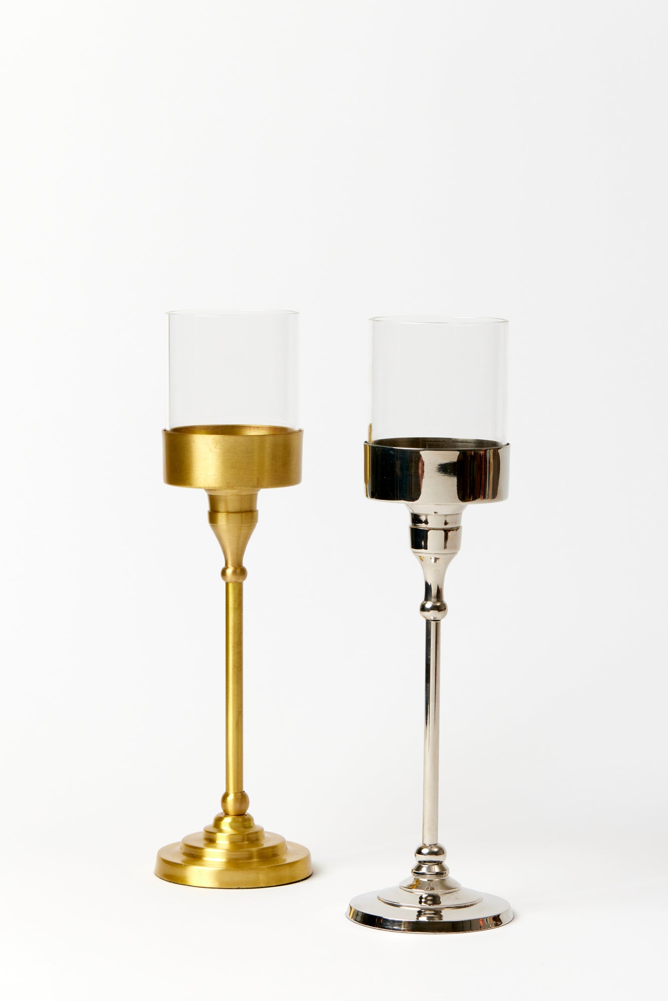 Two table lamp bases with glass containers to hold a tea light and rest the shade atop. Lamp base on the left is brass colored with a brushed finish. Lamp base on the right is nickel silver colored with a polished finish. 
