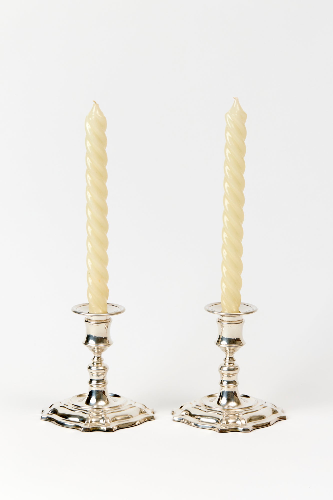 Two candlesticks against a white background. Ivory colored twisty taper candles in polished silver vintage candlestick holders. 
