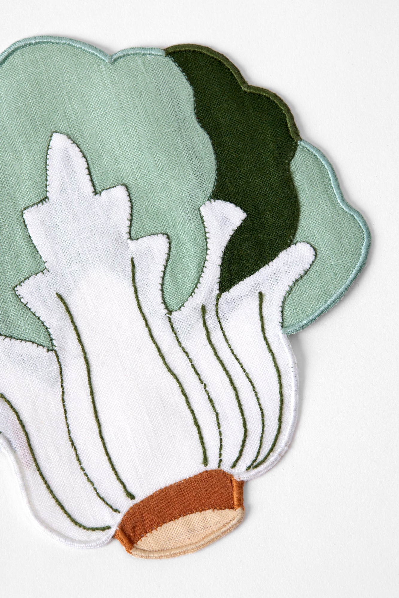 Linen cocktail napkin in the shape of a head of bok choy. Napkin is white center with green leaves and a brown base, along with embroidered details.