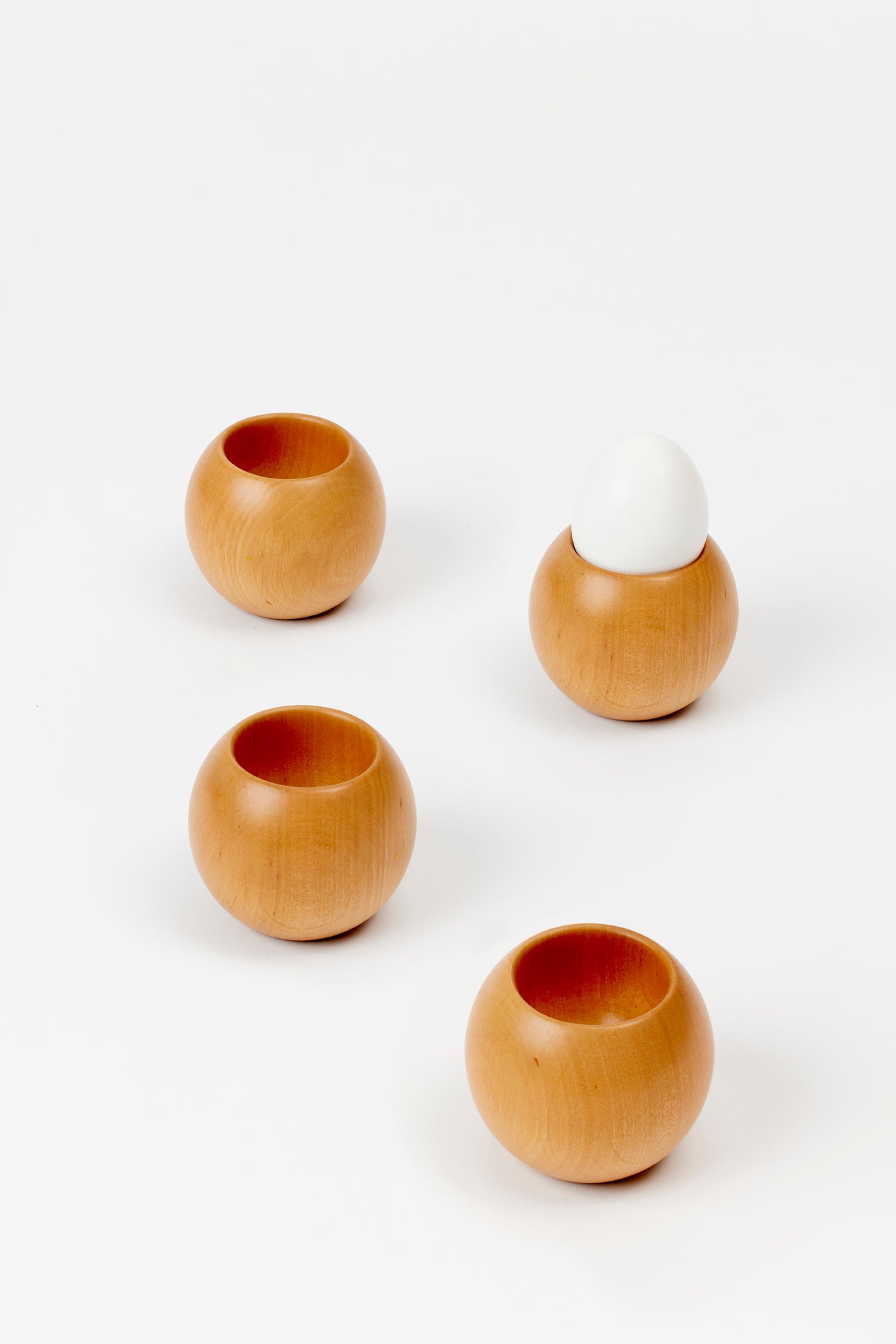 4 round wooden egg cups on a white background. One cup is holding a white egg. Wood is a blonde color. 