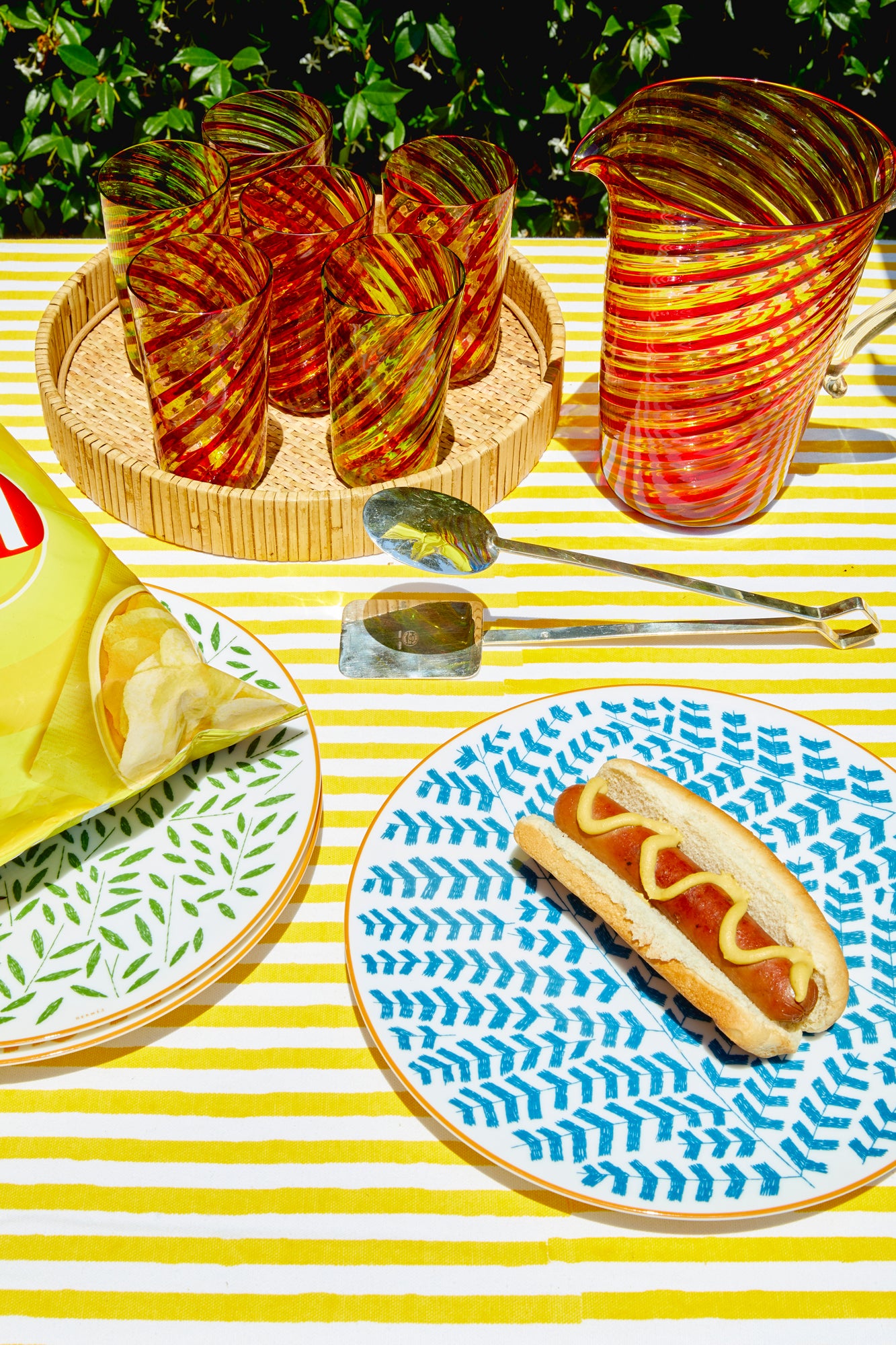 Photograph of outdoor table setting featuring large pitcher and set of tumblers in twisted and colorful Murano glass.