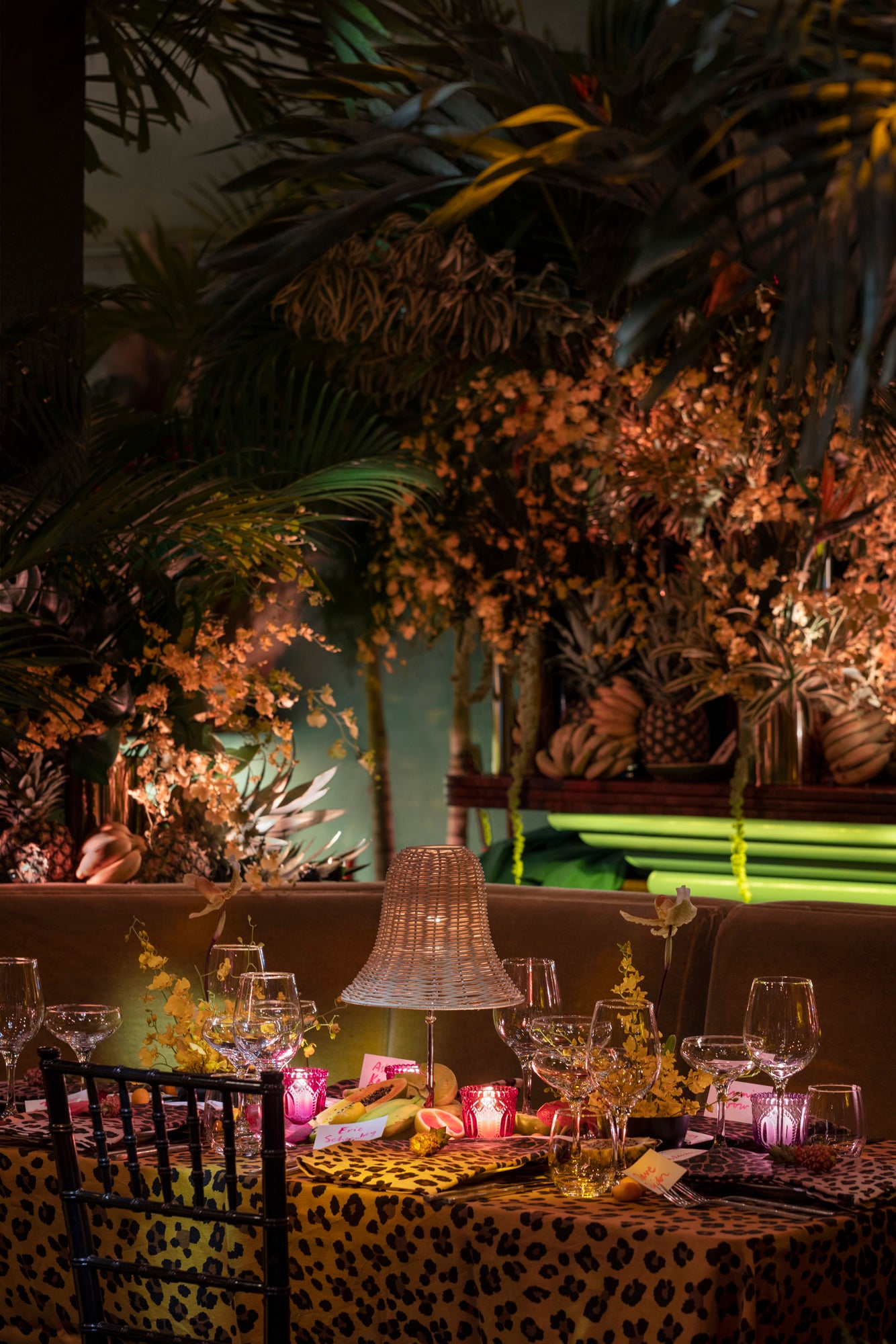 Table setting in a tropical lounge. Setting includes leopard tablecloth, fine dishes and glassware, and a wicker table lamp. Table surrounded by tropical florals. 