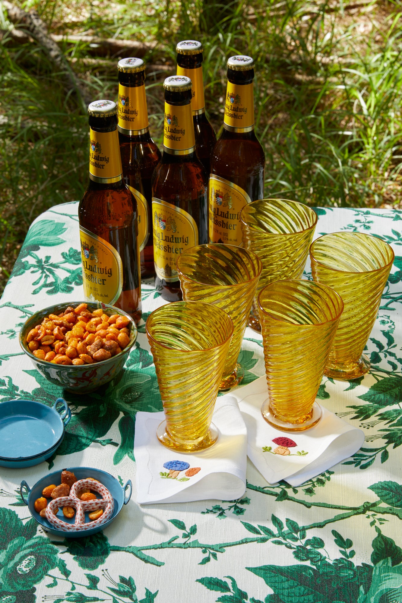 Photograph of table setting featuring French stitch appliqué cocktail napkins, hand blown glass goblets, and a floral tablecloth. 