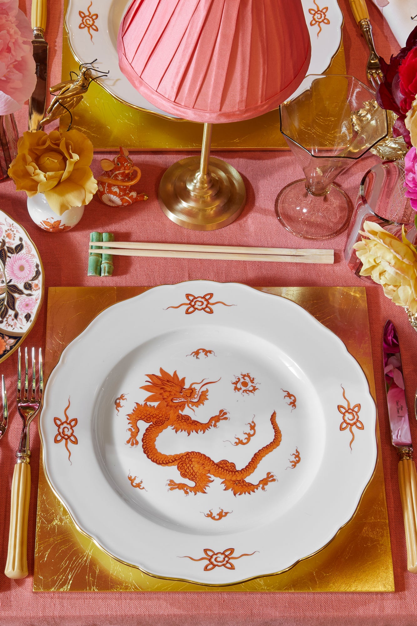 Closeup photograph of table setting featuring square gold placemat, porcelain dragon dinner plate, bamboo utensils, wooden chopsticks on porcelain bamboo rests, hand blown glass goblet, brass table lamp with pink shade, fresh flowers. 